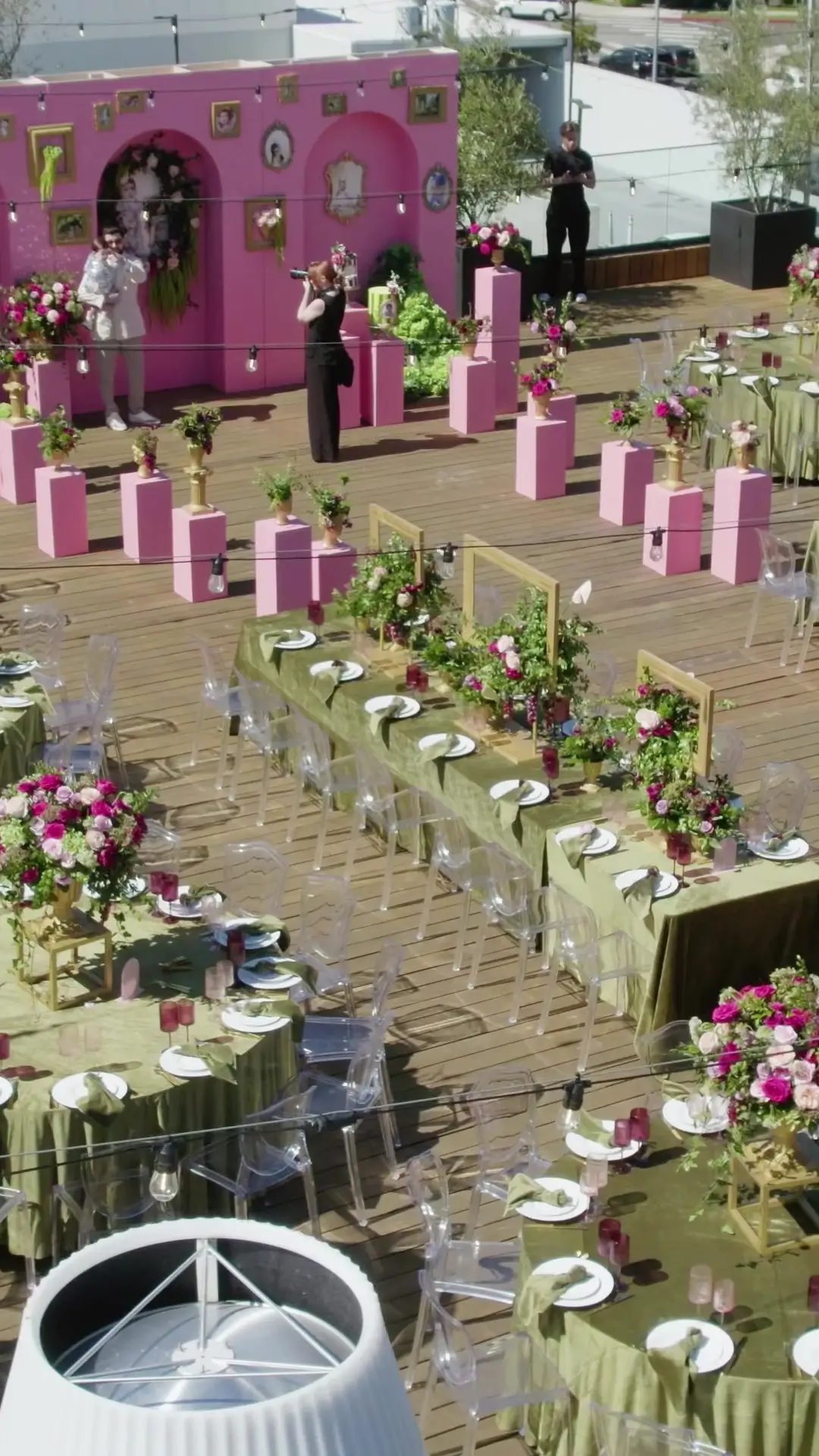 Outdoor event setup with pink stage backdrop and decorative frames, photographers, and string lights over a wooden deck.