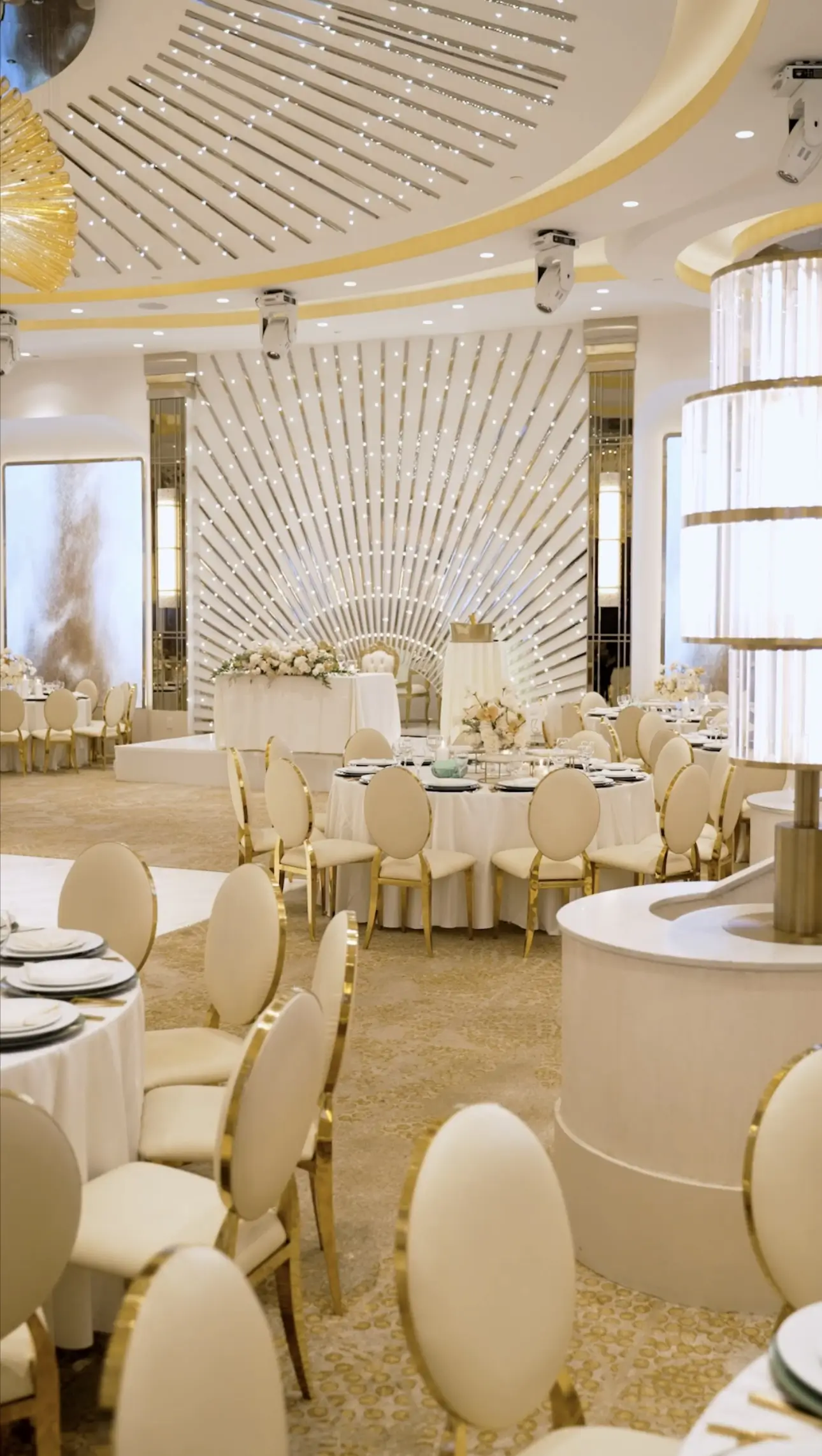 Luxurious banquet hall set for a formal event with round tables, white tablecloths, gold chairs, and a decorative radial backdrop.",